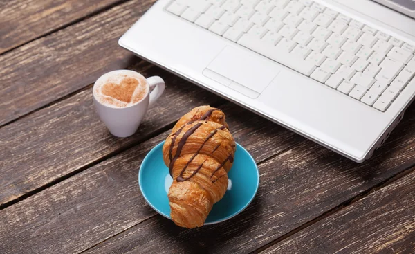 羊角面包和杯咖啡与木制的桌子上的笔记本电脑. — 图库照片