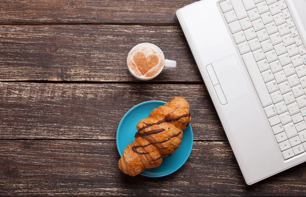 Croissant och en kopp kaffe med laptop på träbord. — Stockfoto