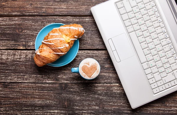 Croissant och en kopp kaffe med laptop på träbord. — Stockfoto