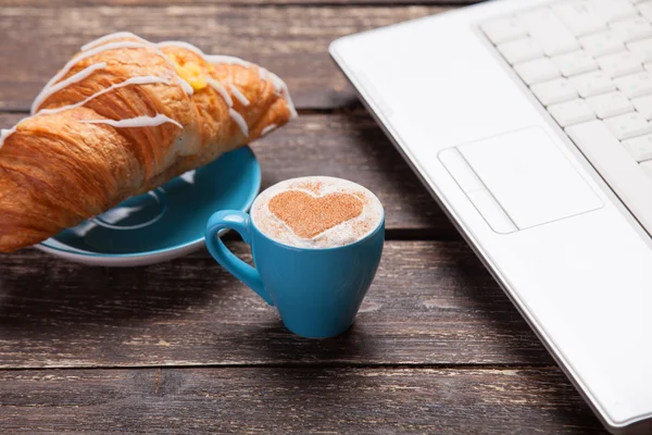 Croissant och en kopp kaffe med laptop på träbord. — Stockfoto