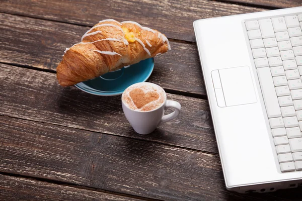 羊角面包和杯咖啡与木制的桌子上的笔记本电脑. — 图库照片