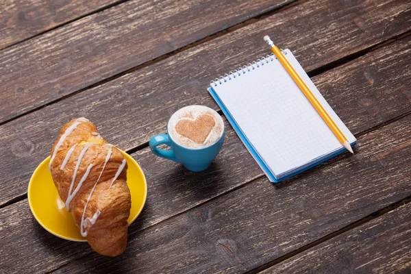 Croissant y taza de café con cuaderno sobre mesa de madera . —  Fotos de Stock