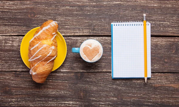 Croissant en koffie met notitieblok op houten tafel. — Stockfoto