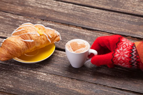 Weibliche Hand im Handschuh hält Tasse Kaffee in der Nähe von Croissant auf Woo — Stockfoto