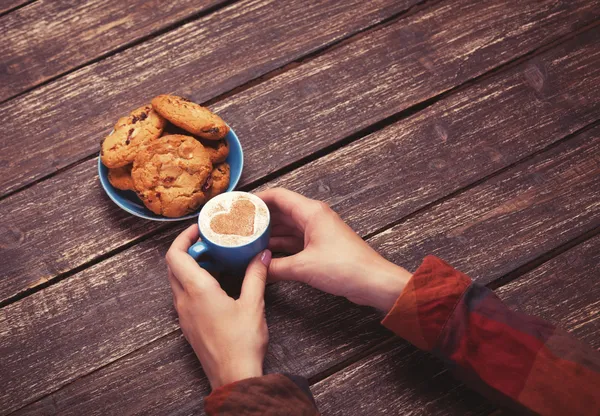 Manos femeninas sosteniendo taza de café y galletas en la mesa de madera . — Foto de Stock