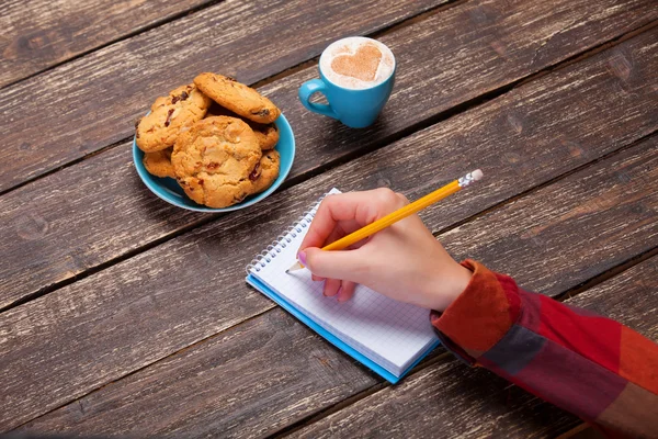 Femme écriture à la main quelque chose dans la note près de cookie et tasse de cof — Photo