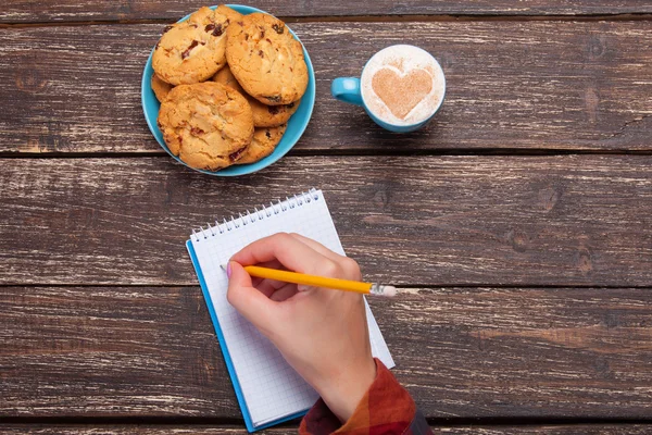 Ręka pisze coś w Uwaga w pobliżu cookie i filiżankę cof — Zdjęcie stockowe