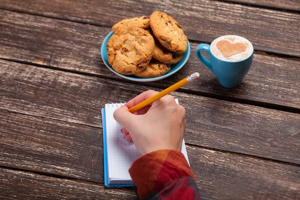 Hembra mano escribiendo algo en la nota cerca de galleta y taza de cof —  Fotos de Stock