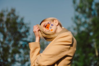 Açık havada yüzünde çiçekler olan kahverengi ceketli sarışın kız.