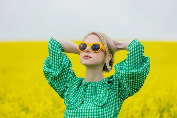 美丽的金发姑娘戴着黄色的太阳镜 穿着绿色的衣服 躺在黄色的菜田里 — 图库照片