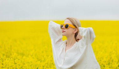 Sarı güneş gözlüklü güzel sarışın ve sarı kolza tohumu tarlasında beyaz gömlekli.