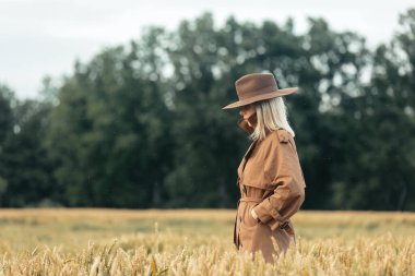 Buğday tarlasında pelerinli ve şapkalı sarışın kadın. 