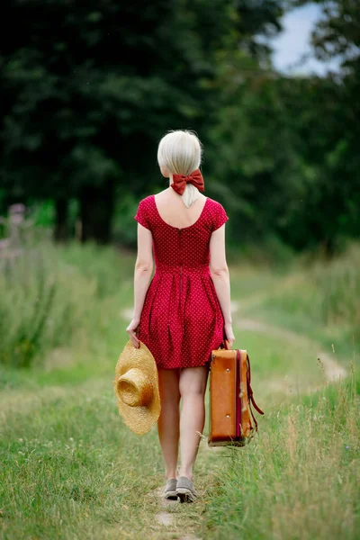 乡村道路上穿着漂亮红色连衣裙 头戴手提箱 头戴帽子的金发女士 — 图库照片