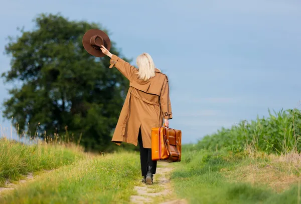 ブロンド女性でマントと帽子でスーツケースは村の田舎道を歩く — ストック写真