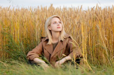 Pelerinli sarışın kadın buğday tarlasının yanında oturuyor. 