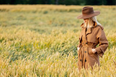 Buğday tarlasında pelerinli ve şapkalı sarışın kadın. 