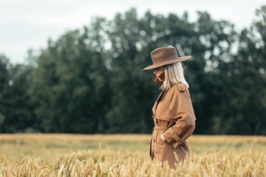 Buğday tarlasında pelerinli ve şapkalı sarışın kadın. 