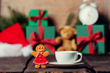 Bir fincan kahve ve kurabiye oyuncağı ahşap masada, arka planda hediyeler var.