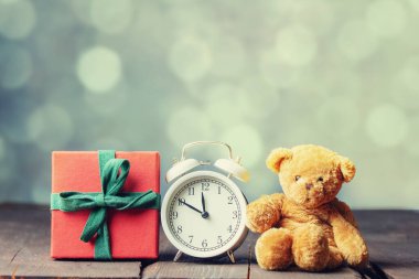 Hediye kutusu ve oyuncak ayı, arka planda bokeh olan ahşap masada çalar saat.