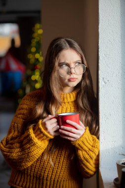 Gözlüklü güzel bir kız ve kupalı sarı bir süveter. Pencerenin yanında ve arka planda Noel ağacı var.