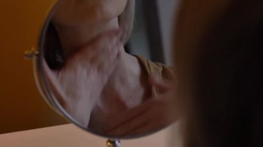 Orta yaşlı kadın aynanın yanındaki masada oturuyor ve yüz masajı yapıyor.