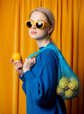 Sarı güneş gözlüklü ve mavi süveterli şık Ukraynalı kadın. Perde arkası ağ çantalı limon.