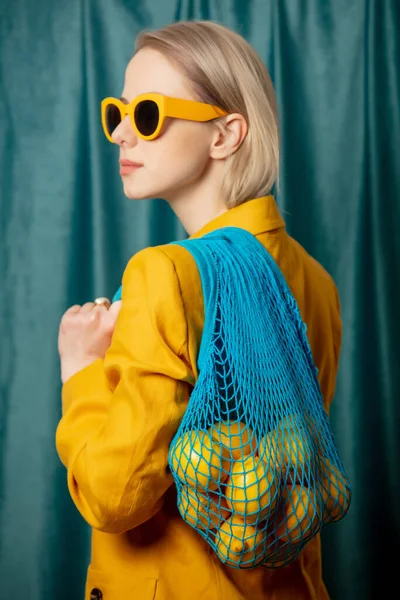 黄色のサングラスと緑のカーテンの背景にネットバッグにレモンとジャケットのスタイリッシュなウクライナの女性 — ストック写真