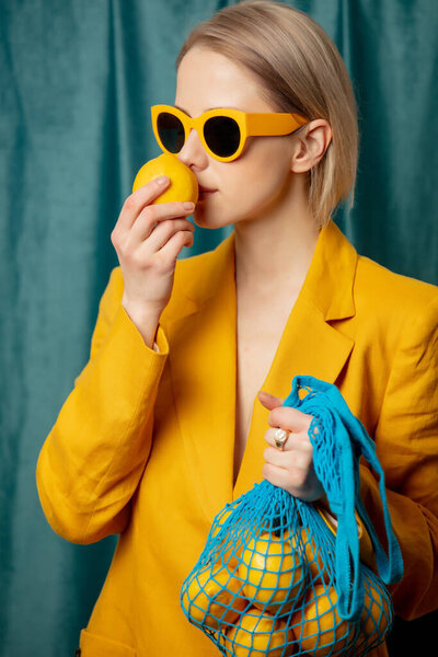 Stylish ukrainian woman in yellow sunglasses and jacket with lemons in net bag on green curtains background