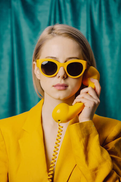 Stylish ukrainian woman in yellow sunglasses and jacket with deal phone on green curtains background