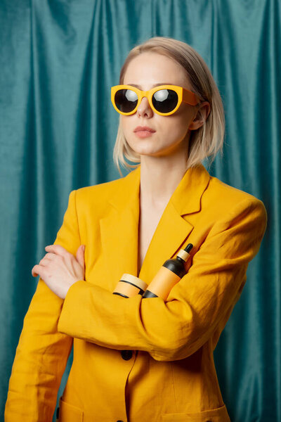 Stylish ukrainian woman in yellow sunglasses and jacket with cosmetic bottle on green curtains background