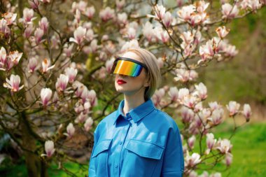 Stylish woman in glasses and blue shirt and VR glasses near Magnolia tree in spring time