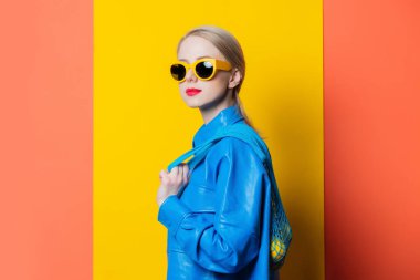 Stylish woman in blue shirt and yellow sunglasses with lemons in net bag on yellow and coral color background
