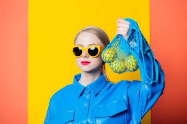 Stylish woman in blue shirt and yellow sunglasses with lemons in net bag on yellow and coral color background