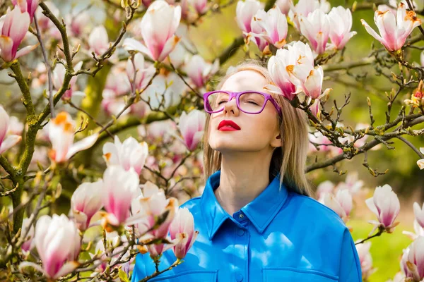 春の時間にマグノリアの木の近くの眼鏡と青いシャツのスタイリッシュな女性 — ストック写真