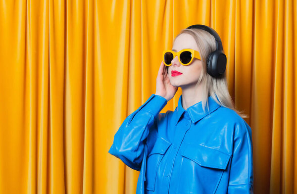 Stylish woman in blue shirt and yellow sunglasses with music headphones against yellow curtain background