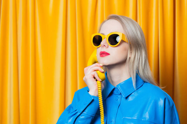 Stylish woman in blue shirt and yellow sunglasses stands with retro dial phone on yellow curtains background