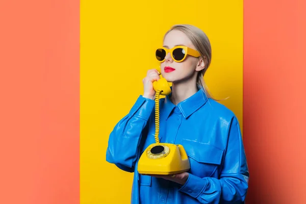 黄色の背景にダイヤル電話で青のシャツと黄色のサングラスでスタイリッシュな女性 — ストック写真