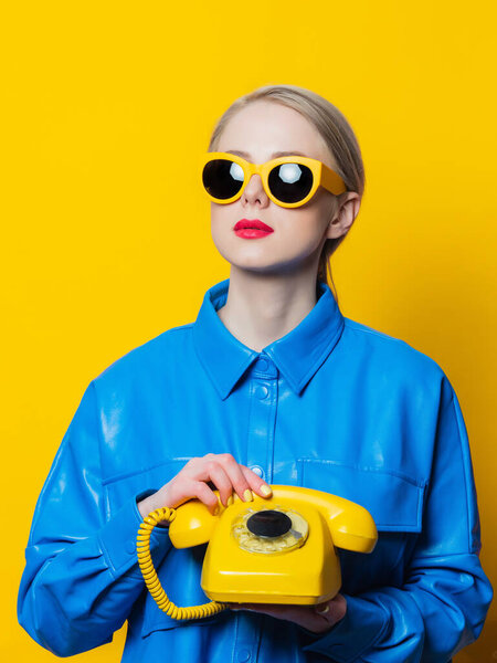 Stylish woman in blue shirt and yellow sunglasses with dial phone on yellow background
