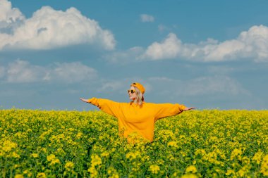 Stylish woman in yellow hoodie, cap and sunglasses in rapeseed field