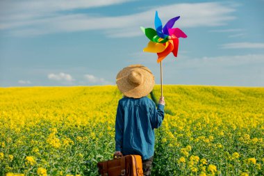 Şapkalı, bavullu ve kolza tarlasında fırıldak taşıyan bir kadın.