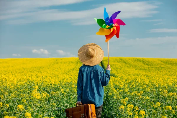 Şapkalı, bavullu ve kolza tarlasında fırıldak taşıyan bir kadın.