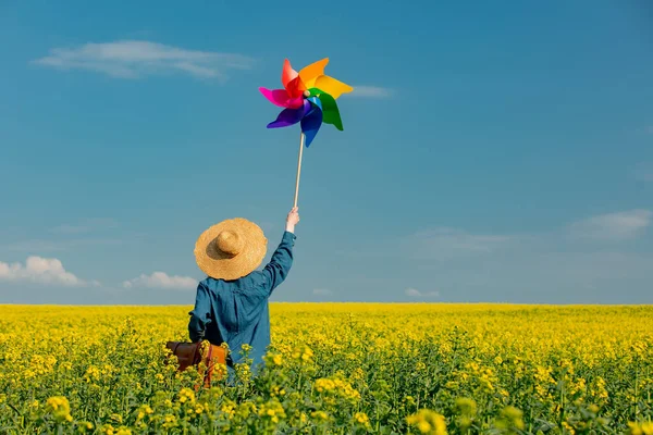 Şapkalı, bavullu ve kolza tarlasında fırıldak taşıyan bir kadın.