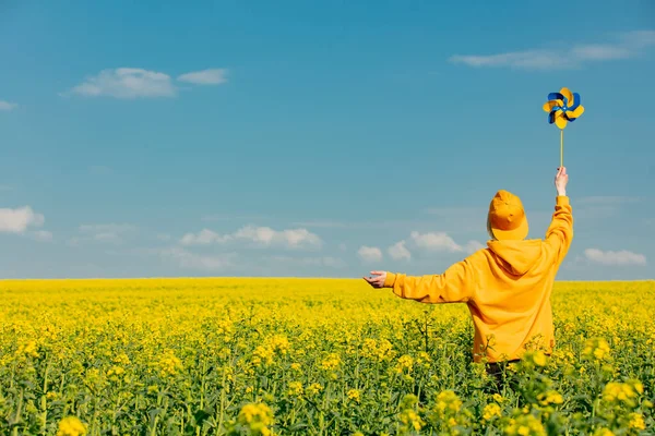 Kapüşonlu ve şapkalı bir kadın, kolza tarlasında fırıldak gibi. 