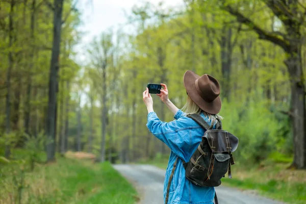 Şapkalı ve sırt çantalı şık bir kadın açık havada cep telefonuyla fotoğraf çeker.