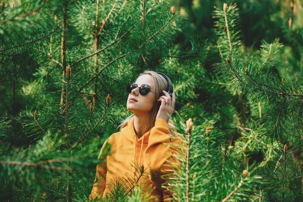 穿着连帽衫的时髦女孩在松树林的耳机里听音乐 — 图库照片