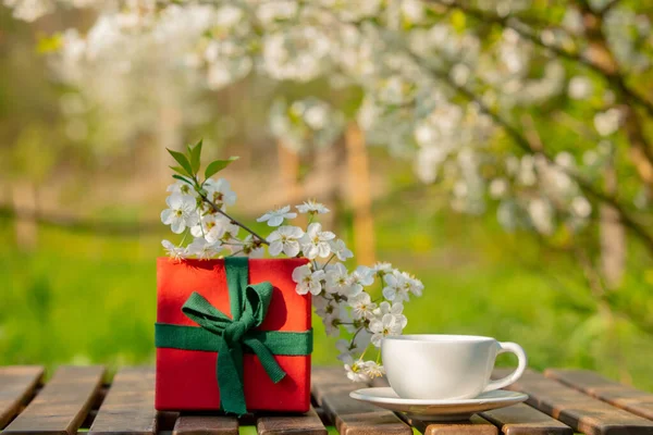 Cup of coffee and gift box on wooden table in blooming tree garden