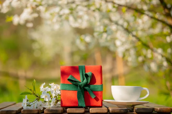 Cup of coffee and gift box on wooden table in blooming tree garden