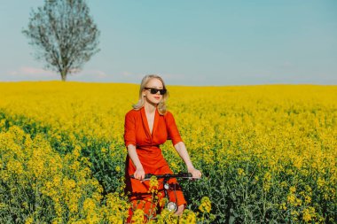 Klasik elbiseli, güneş gözlüklü, kolza tohumu tarlasında bisiklet süren güzel sarışın kız.