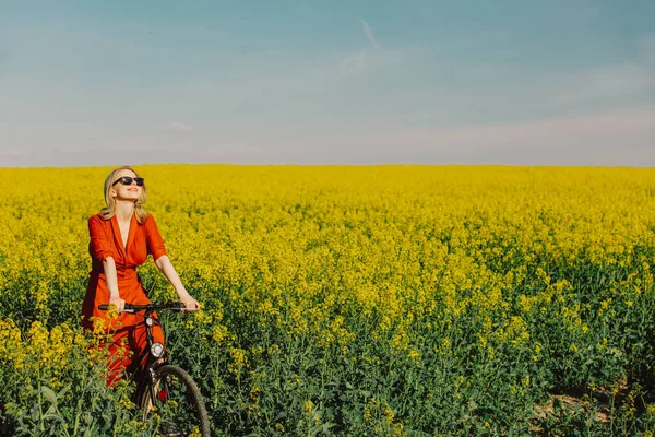 春天的时候 美丽的金发姑娘穿着老式服装 戴着墨镜 骑自行车在菜田里 — 图库照片