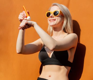 Bikinili kız vücuduna turuncu arka planda güneş kremi sürüyor.